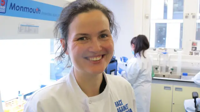 A cientista Claire Durrant sorri. Ela tem o cabelo preso e olha para a câmera em seu laboratório. Ela está vestindo um jaleco branco com as palavras “Race Against” claramente escritas e a palavra demência pode ser inferida. Há pessoas trabalhando atrás dela.