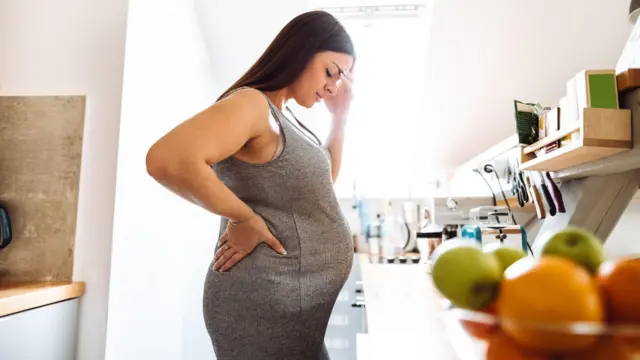 De pé em frente a um balcão na cozinha, mulher grávida, com longos cabelos castanhos e vestido cinza, segura as costas com a mão direita e a cabeça comm a esquerda. Ao fundo, maçãs verdes e laranjas em uma cesta de frutas.