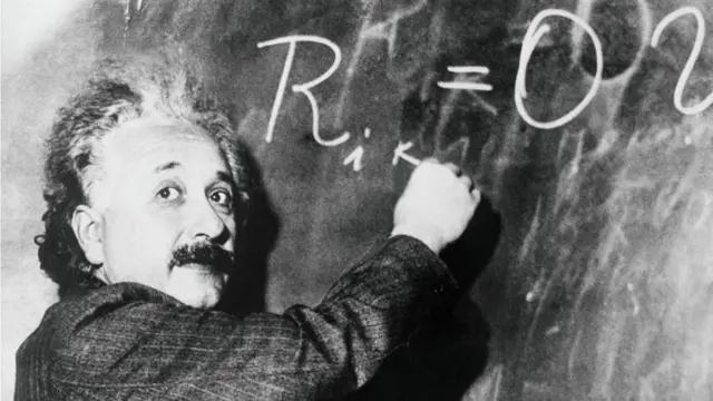 Albert Einstein olhando para a câmera enquanto escreve uma fórmula na lousa