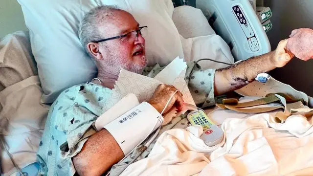 Peter Smith está deitado em uma cama de hospital, estendendo a mão para tocar a mão de outra pessoa (não retratada). Ele usa óculos e uma camisola hospitalar, cercado por equipamentos médicos
