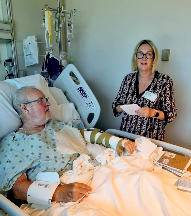 Peter Smith com o braço esquerdo enfaixado, deitado em uma cama de hospital, com a mulher, Joanna, ao seu lado