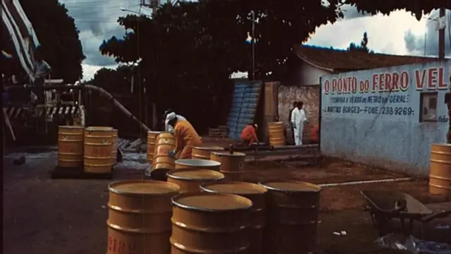 Área externa de um ferro-velho com vários tambores metálicos amarelos espalhados pelo chão, enquanto pessoas usam roupas de proteção para manuseá-los; ao fundo, há um muro com a inscrição “O Ponto do Ferro Velho” e árvores ao redor, em uma operação de descontaminação.