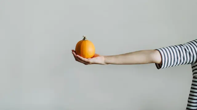 Braço esticado de uma pessoa de blusa listrada segura uma fruta na mão