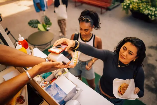 Mulher está pagando com celular por hamburguer que comprou em carrinho de comida. Ela aproxima o aparelho de uma maquininha de cartão.