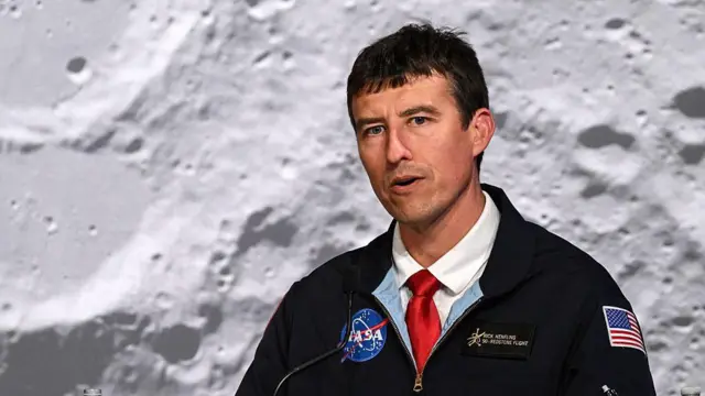 Homem de meia-idade com uniforme da Nasa e gravata vermelha fala em frente a uma enorme fotografia da superfície lunar