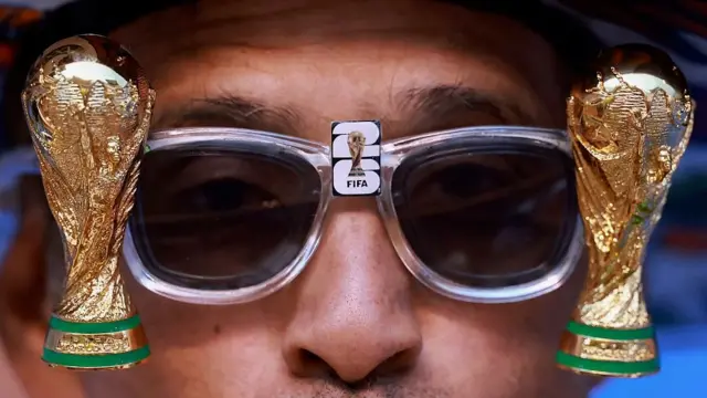 Uma foto em close-up de um homem usando óculos escuros com uma miniatura da Copa do Mundo em cada lado.