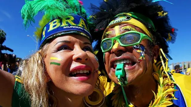 Uma foto em close-up de dois torcedores brasileiros vestindo roupas verdes e amarelas, além de cocares de penas.