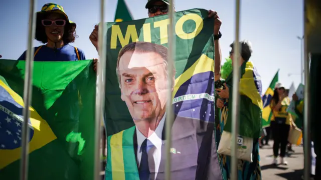 Manifestantes seguram bandeiras do Brasil e um grande estandarte com a imagem do ex-presidente Jair Bolsonaro durante manifestação. A cena é vista através de grades metálicas em primeiro plano, enquanto participantes vestindo roupas nas cores verde e amarelo se concentram em uma área aberta sob a luz do dia.