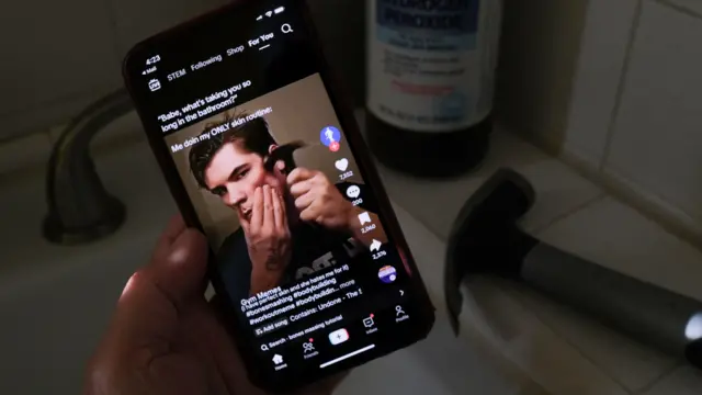 Um homem segura um smartphone exibindo um vídeo mostrando um jovem diante da câmera em um conteúdo relacionado a rotina de cuidados pessoais. A cena ocorre em um banheiro, identificado pela pia, torneira e pelos objetos ao redor, incluindo um frasco ao fundo e um aparelho elétrico sobre a bancada. A iluminação é baixa e concentra a atenção na tela do celular.