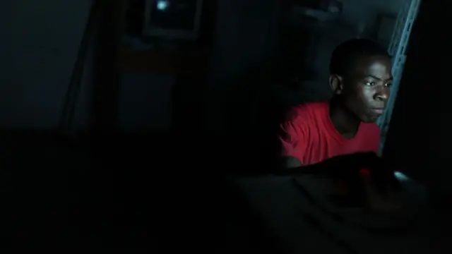Um jovem aparece sentado diante de uma tela em um ambiente escuro, iluminado principalmente pela luz azulada do monitor que destaca seu rosto concentrado. Vestindo uma camiseta vermelha, ele olha fixamente para a tela enquanto utiliza um computador, cercado por um espaço pouco iluminado e com poucos detalhes visíveis ao fundo.