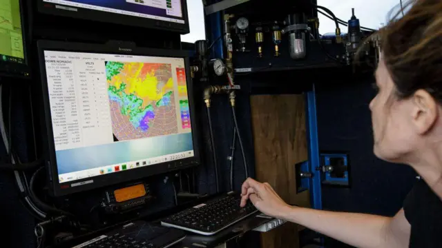 Uma meteorologista monitora uma tempestade a bordo do veículo Doppler on Wheels. Ela veste camiseta preta e observa uma tela de computador com uma imagem colorida