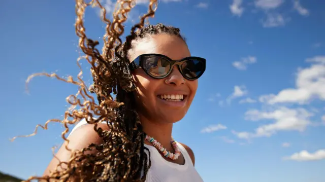 Mulher jovem de óculos escuros e cabelos esvoaçantes em ambiente externo, em dia de sol